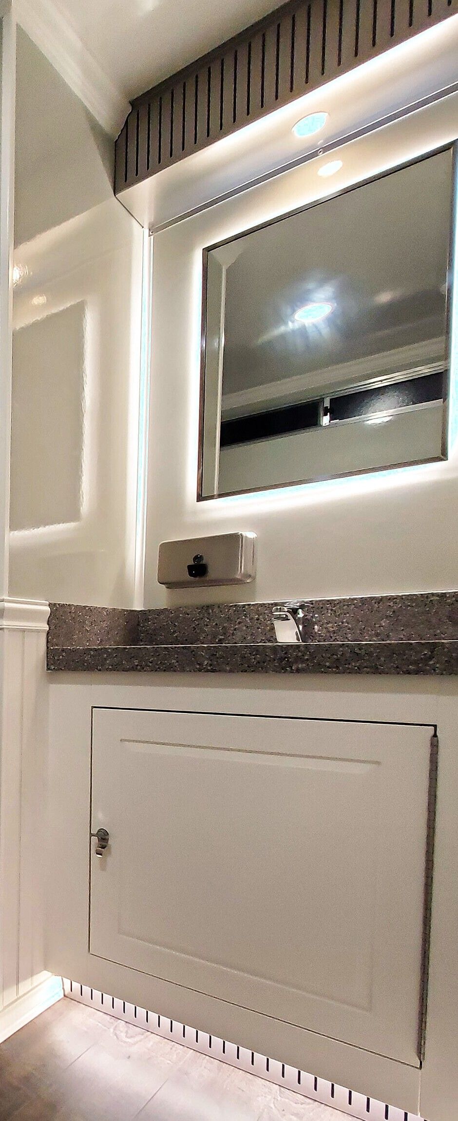A lit mirror over a solid-surface vanity with a faucet and soap dispenser, inside the executive trailer