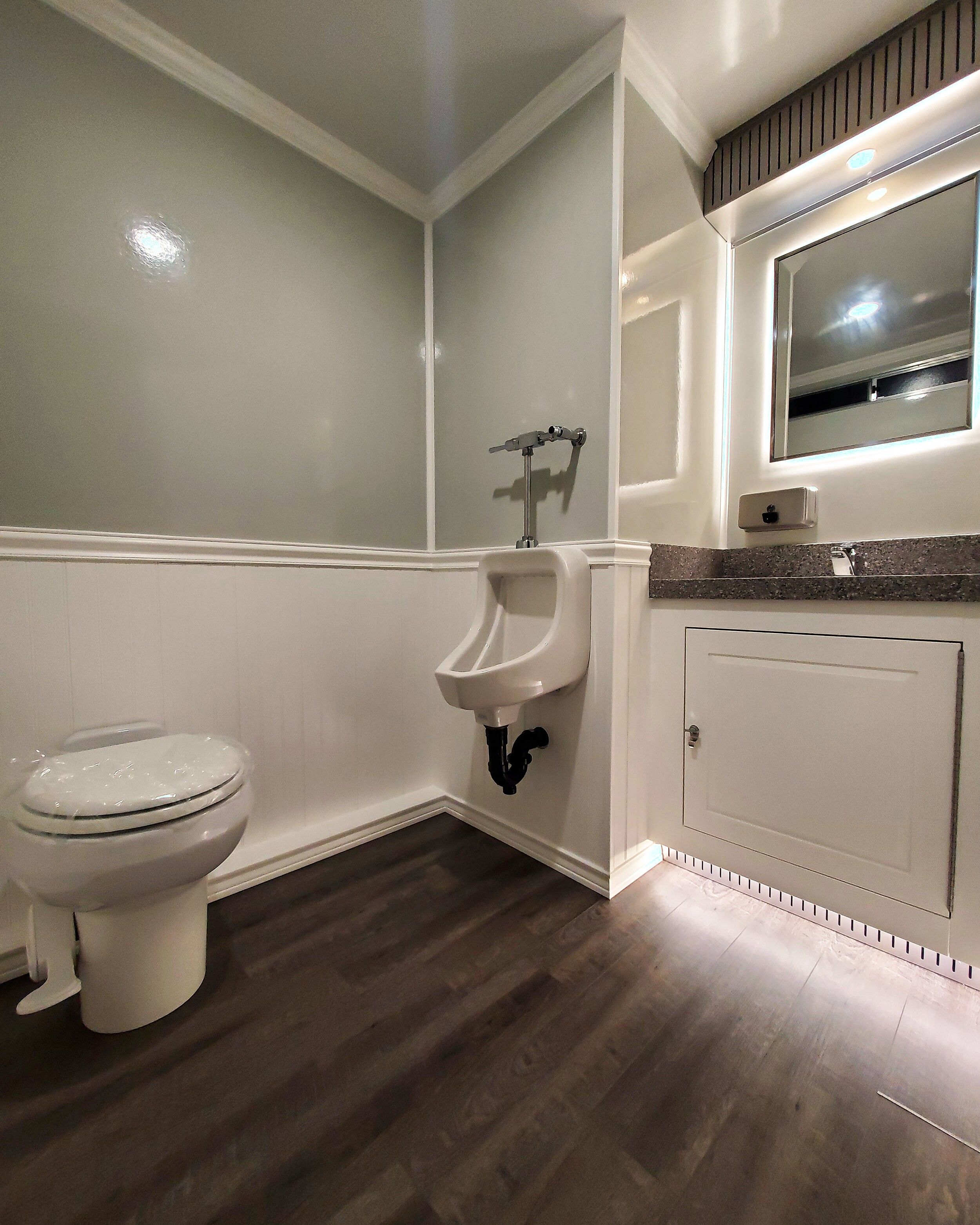 Inside one private room of the executive trailer: a flushing porcelain toilet, a solid-surface vanity under a lit mirror, and vinyl plank flooring