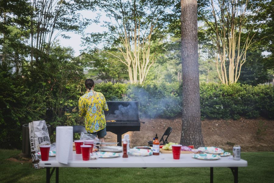 Someone grilling in a leafy backyard beside a folding table set with food and drinks