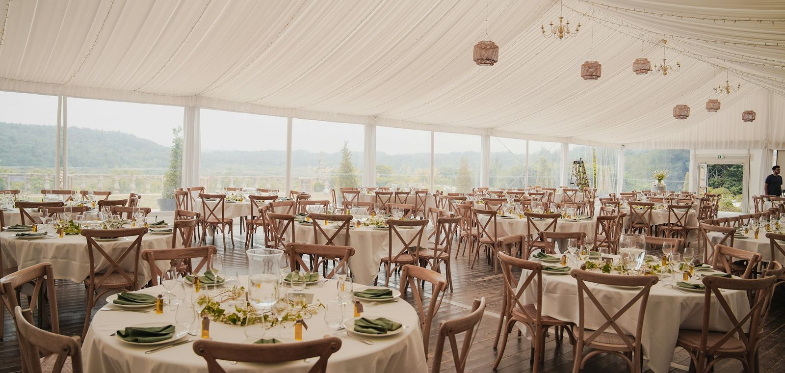 Inside a white wedding tent: round tables set for dinner, wooden chairs, and paper lanterns, with open sides looking out to the hills