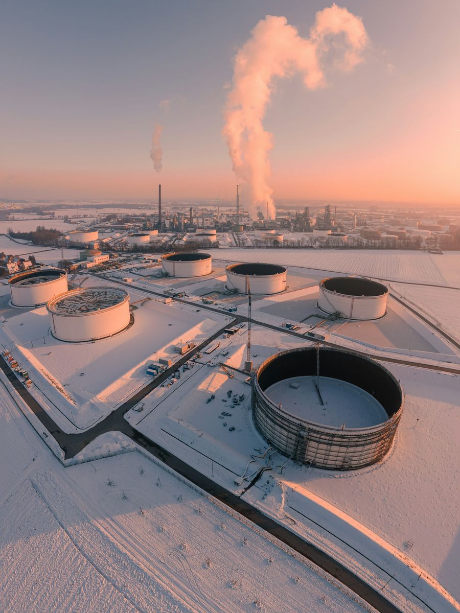 Storage tanks at an industrial site under fresh snow at sunrise, with steam rising into a pink sky